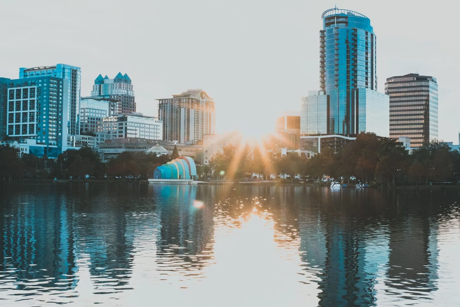 Glass towers reflected in a lake at sunrise, the sun breaking through the trees on the shore