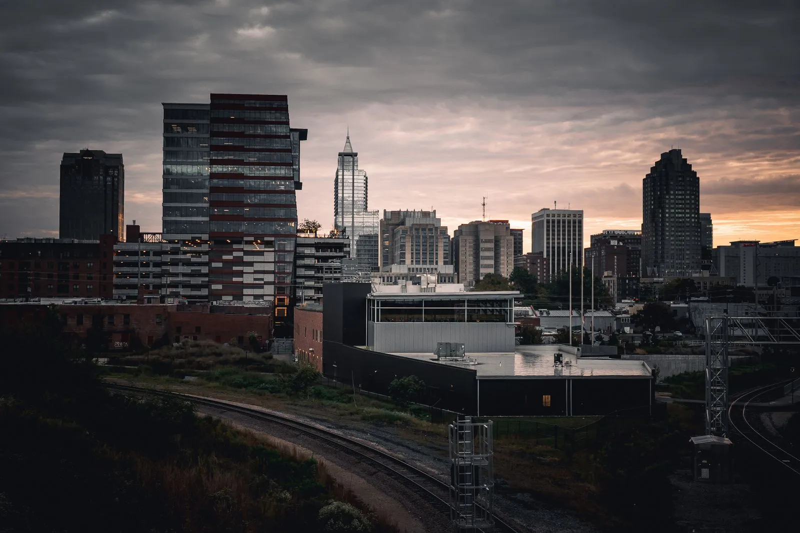 A downtown skyline in silhouette under a stormy sunset sky, rail tracks curving through the foreground
