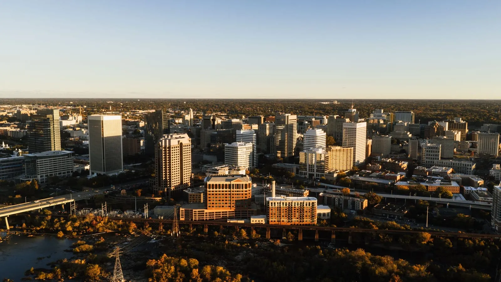 A downtown skyline in warm evening light above autumn trees along a river