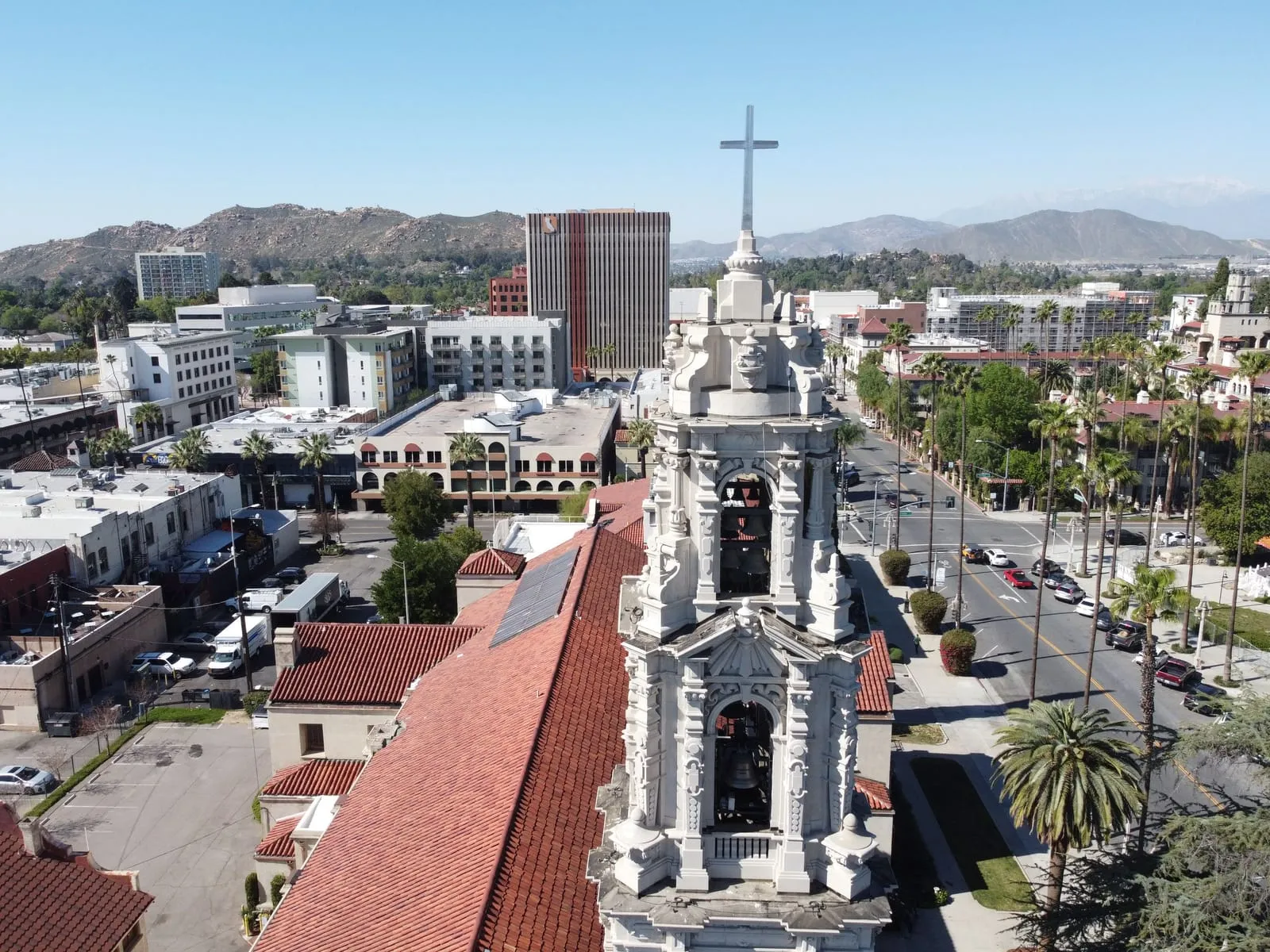 An ornate white bell tower and red tile roofs above palm-lined streets, hills beyond