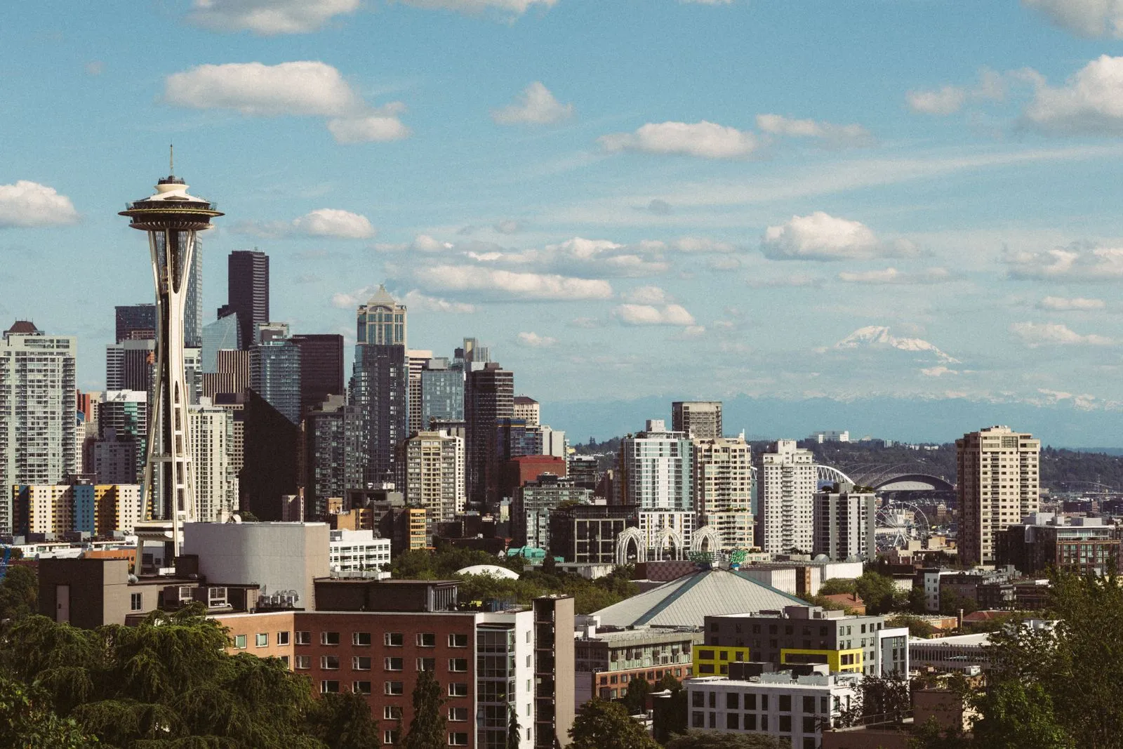 A needle-shaped observation tower in front of a downtown skyline, a snowy peak faint in the distance