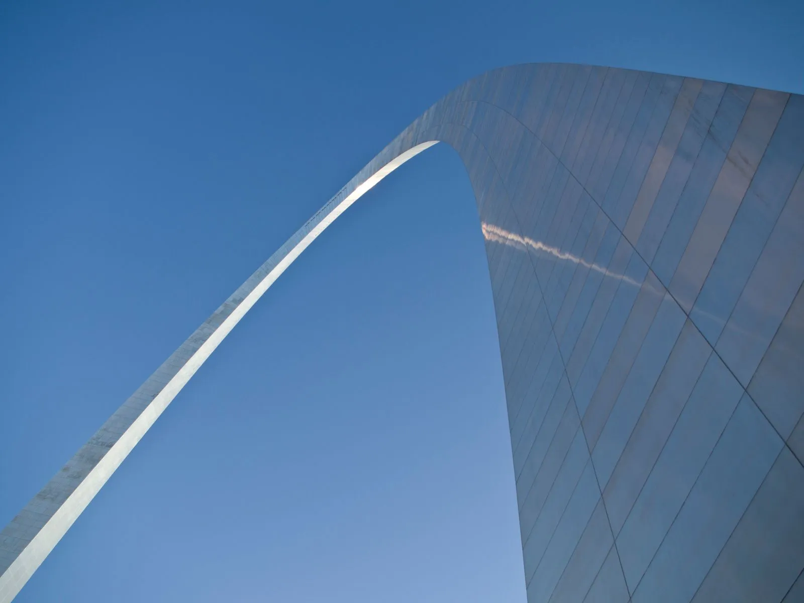 Looking up at a tall curved stainless-steel arch against a deep blue sky