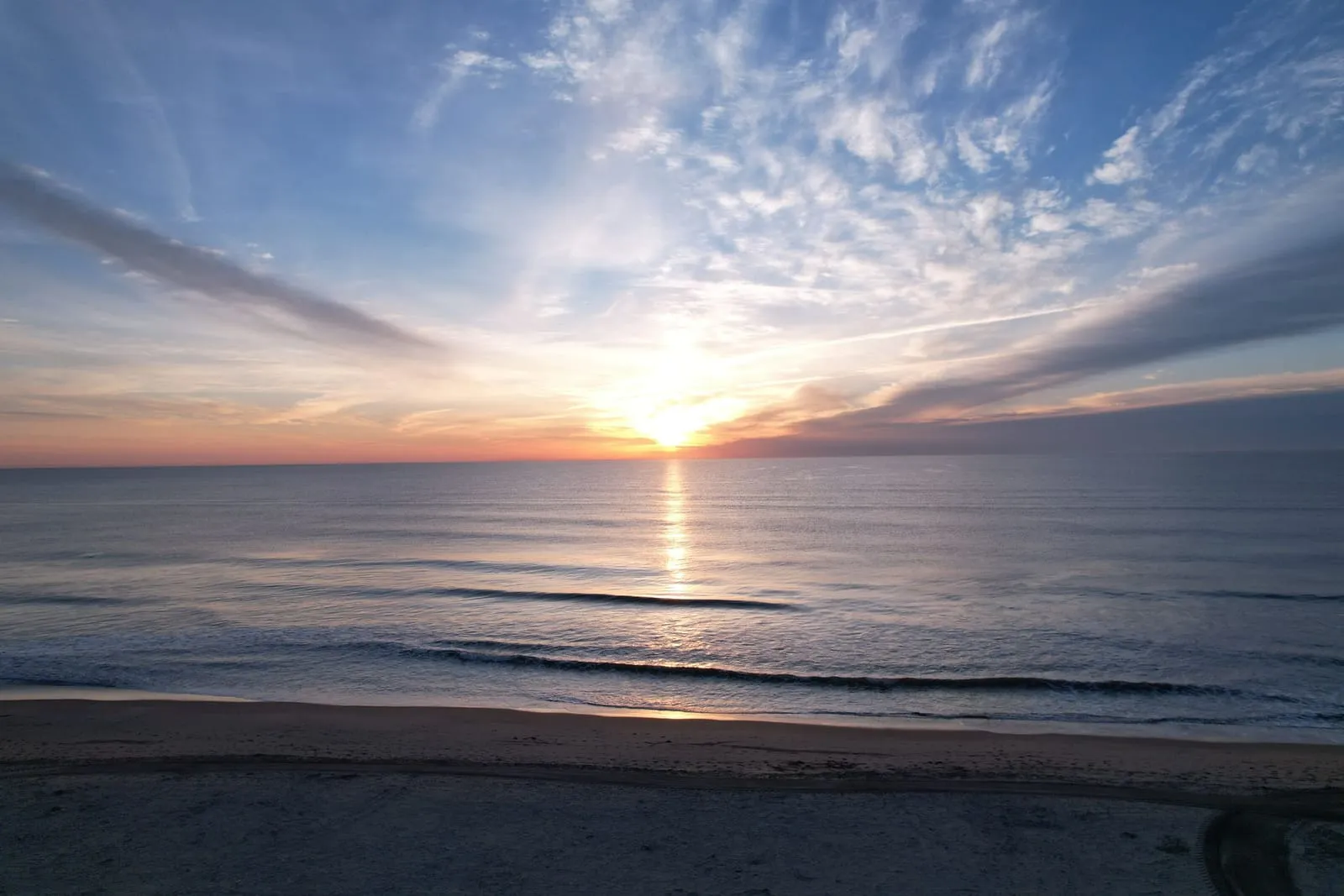 The sun rising over the ocean, streaked clouds above and gentle surf on a wide beach