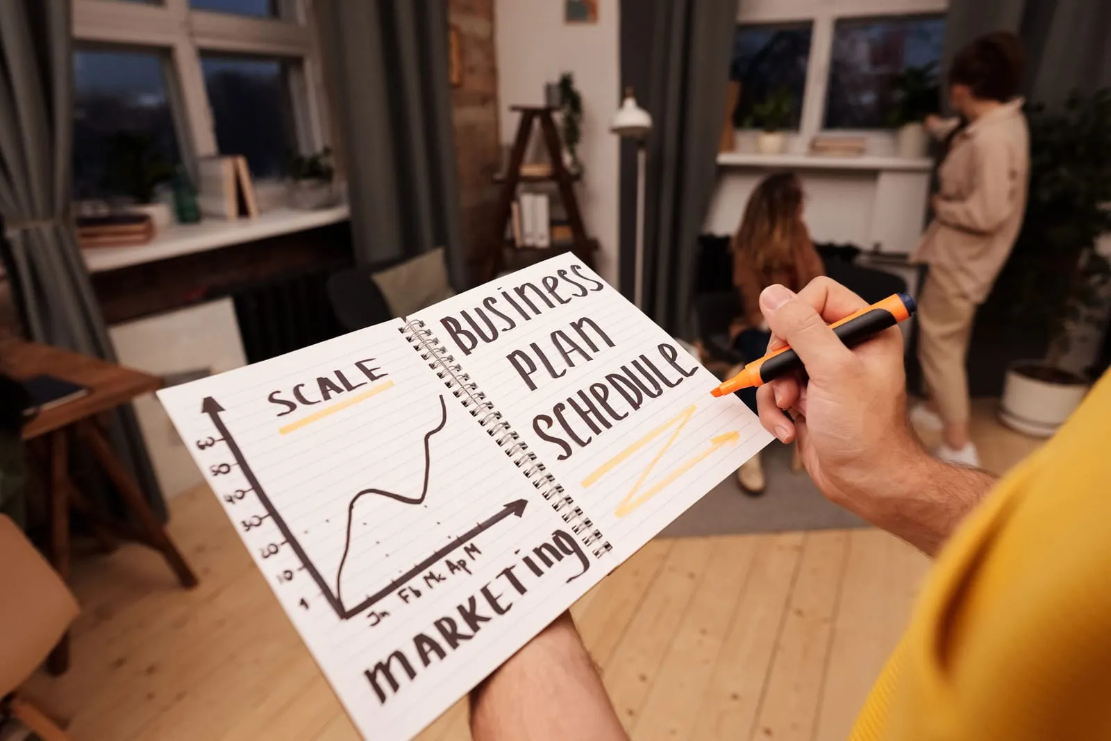 A hand holding a marker over a spiral notebook labeled business plan schedule, with a hand-drawn growth chart and a marketing note, while two people talk in the background.