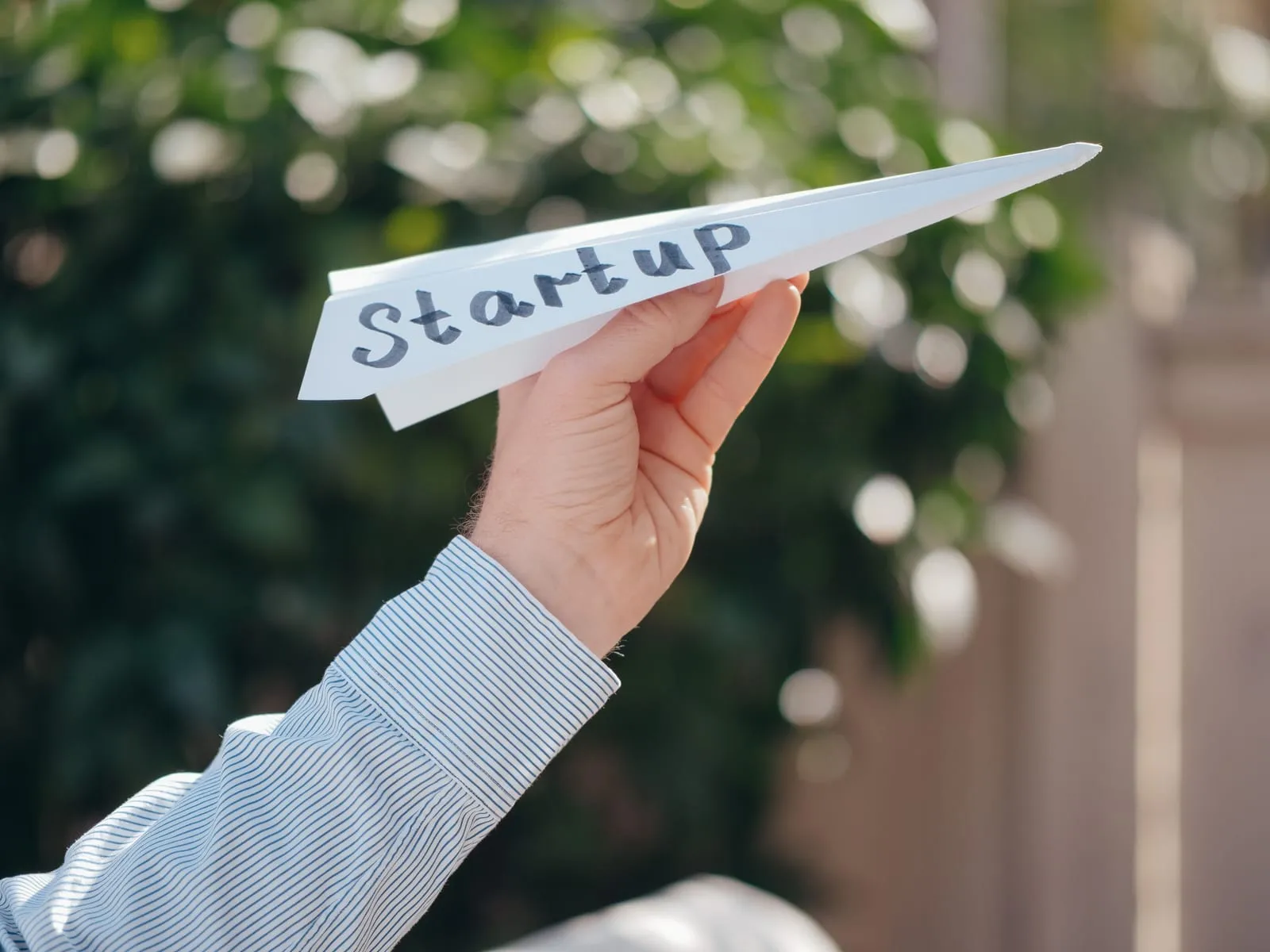 A hand in a striped shirt sleeve holds up a white paper airplane with the word Startup written on it against blurred green foliage.