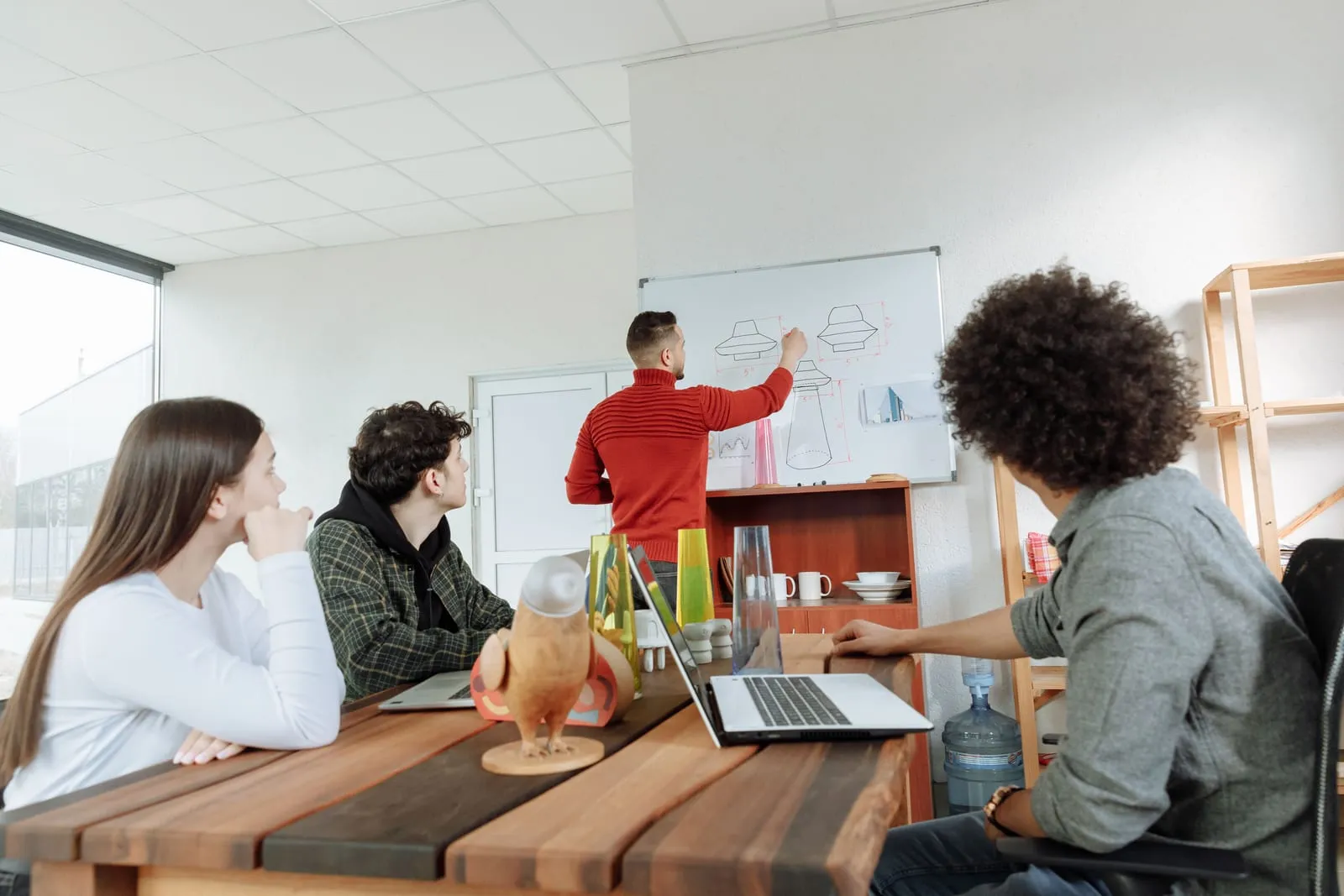 A man in a red sweater sketches product designs on a whiteboard while three people seated at a wooden table with laptops watch him.
