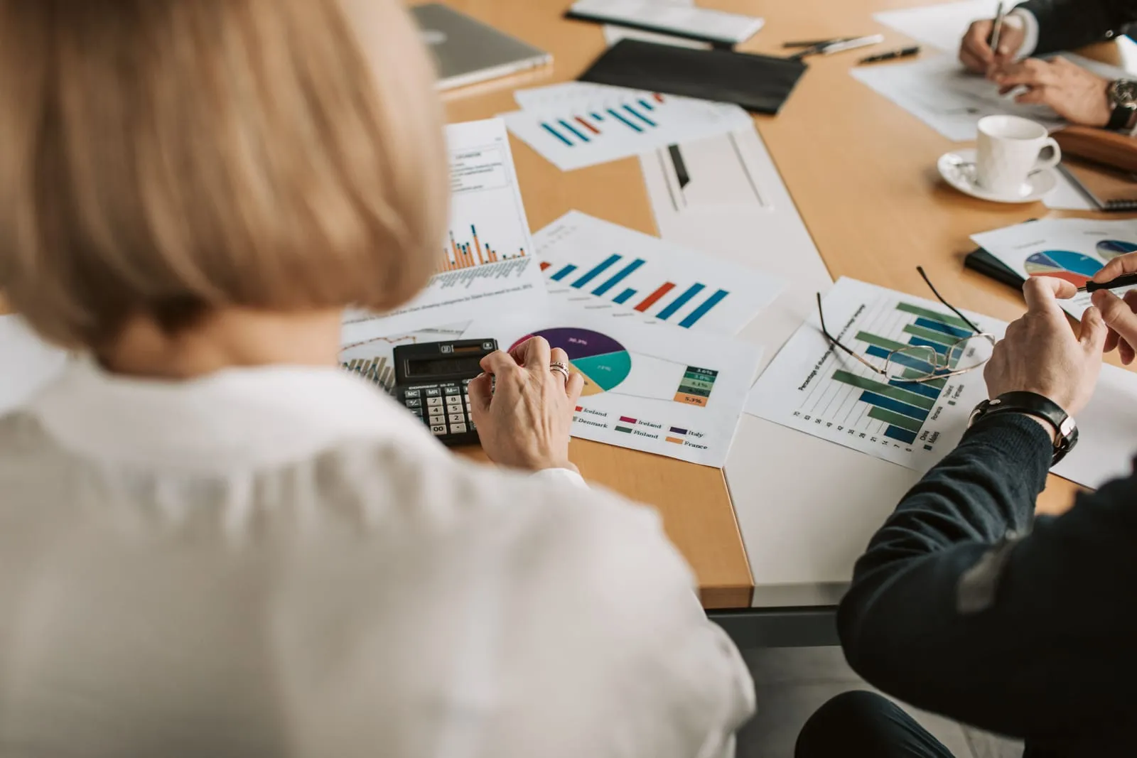 Colleagues around a conference table review printed pie and bar charts, with one person using a calculator and another holding a pen and eyeglasses.