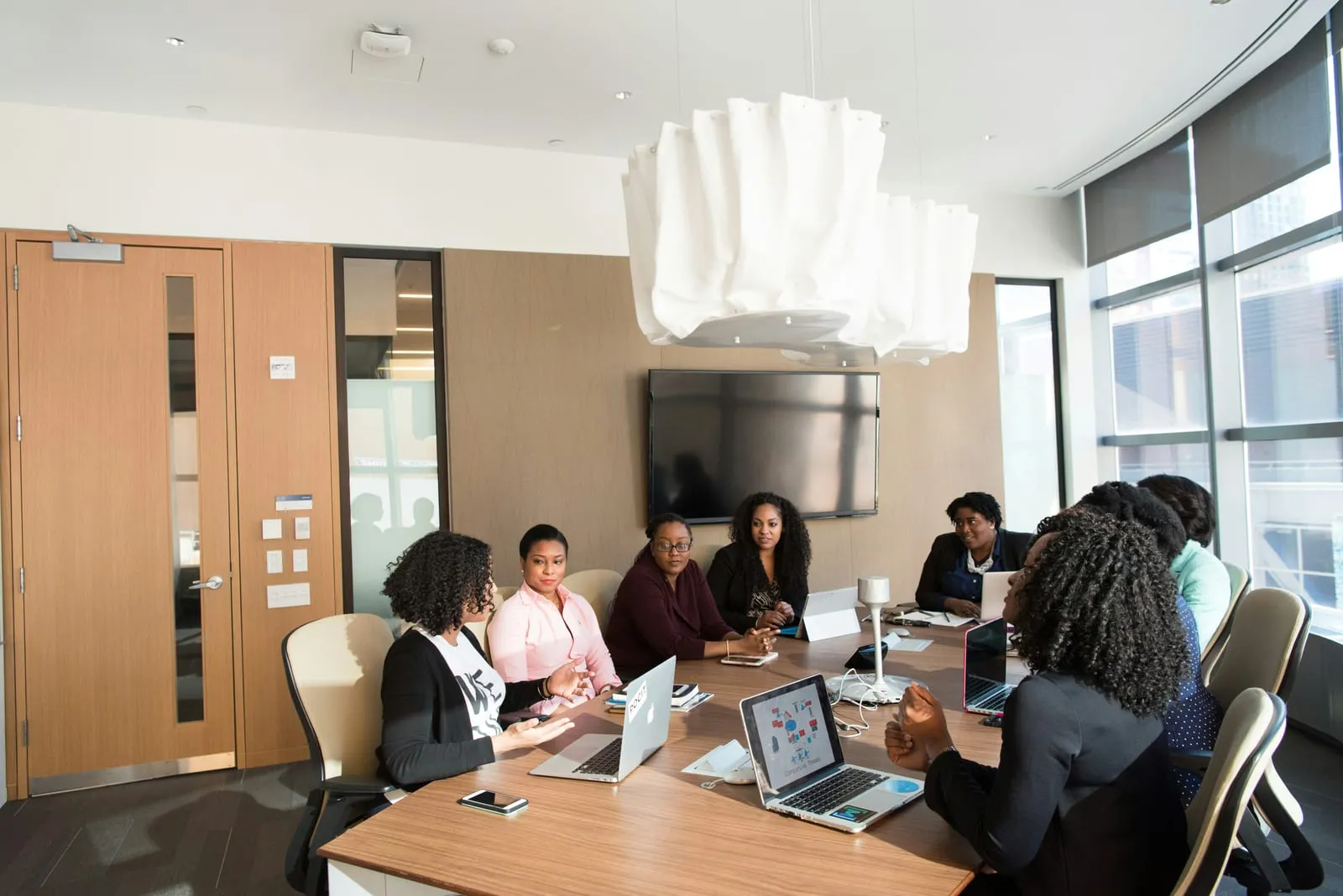 A group of professionals talk around a long wooden table in a bright boardroom with laptops, a wall-mounted screen and city views through large windows.