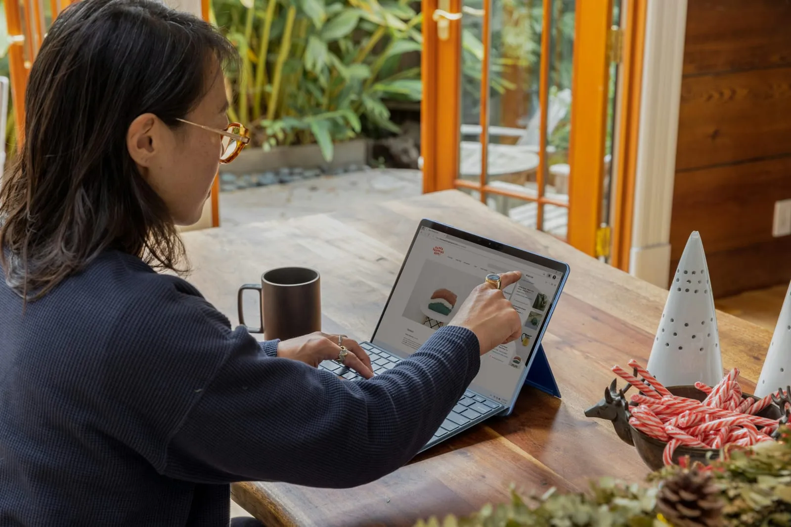 A woman with glasses sits at a wooden table in a sunlit room, touching the screen of a tablet laptop that shows an online shop page.