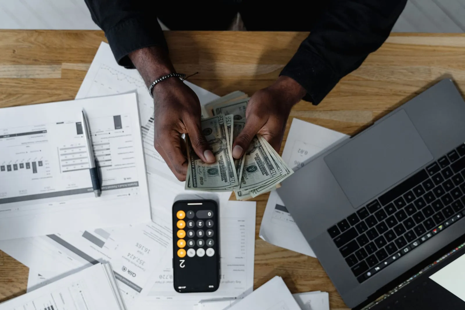 Overhead view of hands counting a fan of twenty-dollar bills beside a smartphone calculator, printed financial reports and an open laptop on a wooden desk.