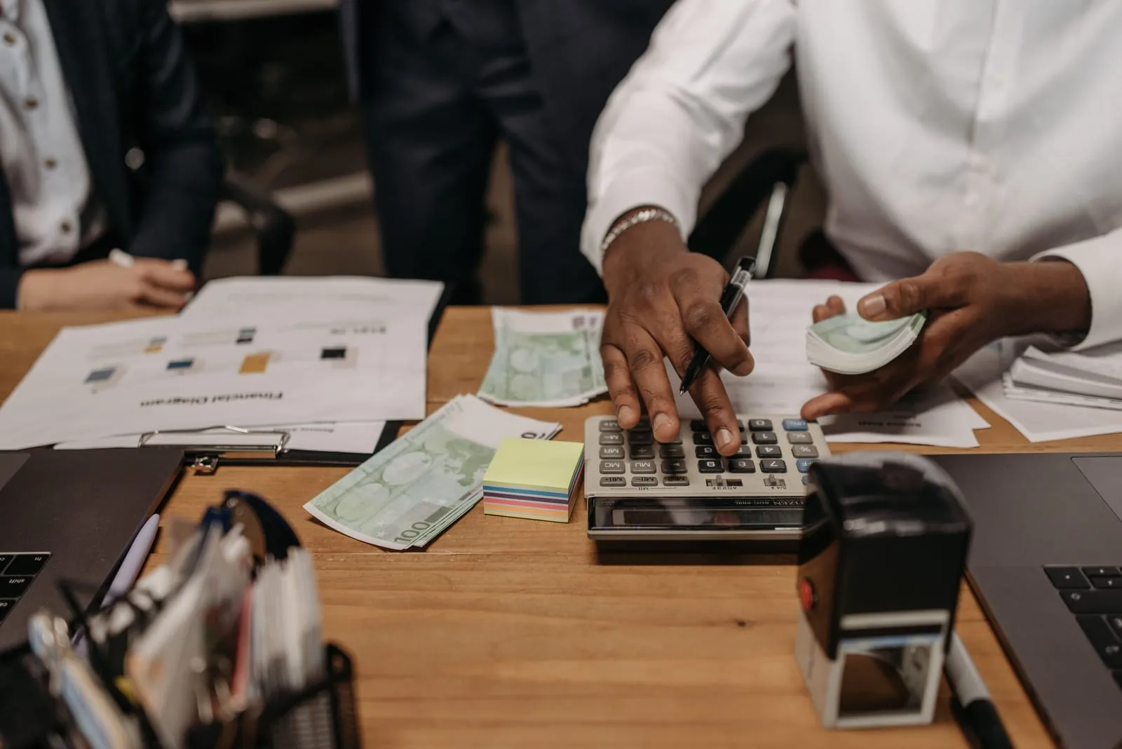 A person in a white shirt taps a desk calculator and holds a stack of euro notes, with more bills, a financial diagram printout and a stamp on the desk.