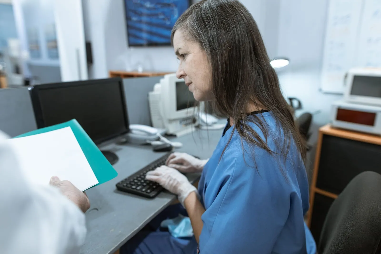 A woman in blue scrubs and gloves types at a computer in a clinic office while a colleague hands her a teal folder with a document.