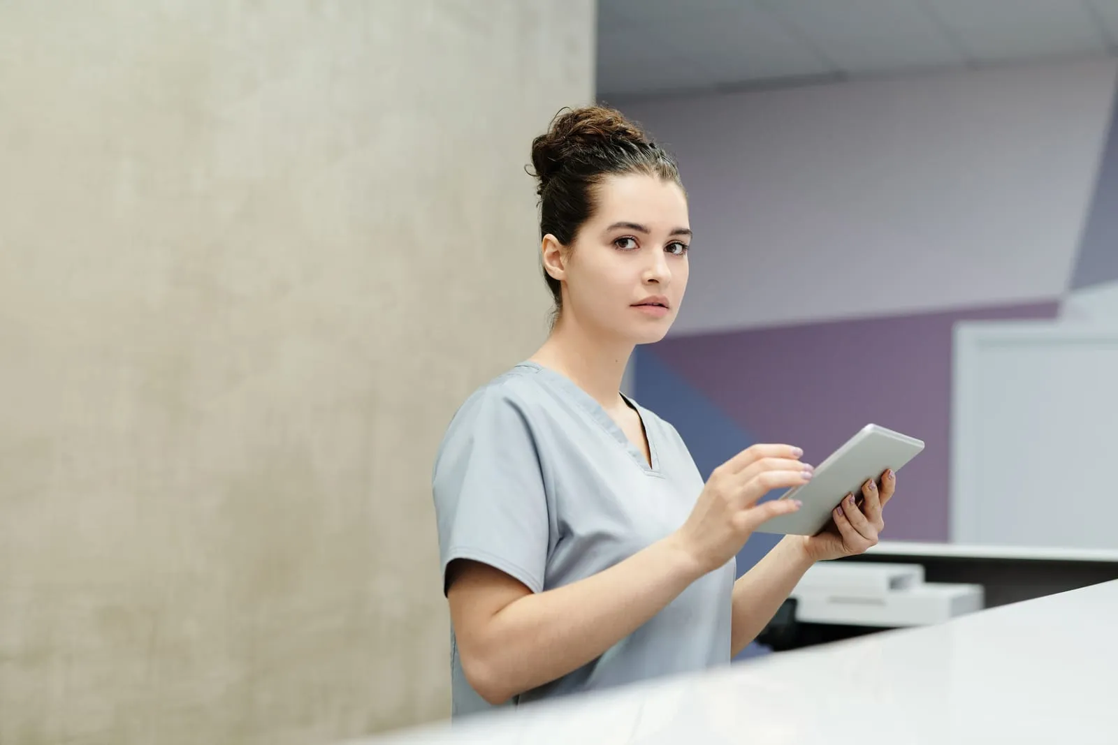 A healthcare worker in gray scrubs stands at a reception counter holding a tablet and looking toward the camera.
