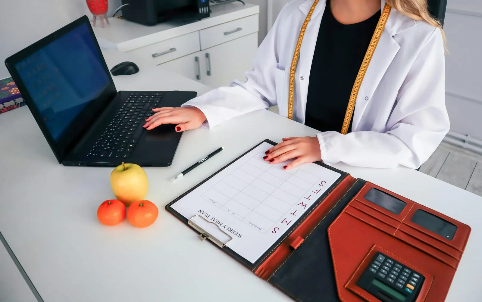 A woman in a white coat with a tape measure around her neck works on a laptop beside a weekly meal plan clipboard, fruit and a calculator.