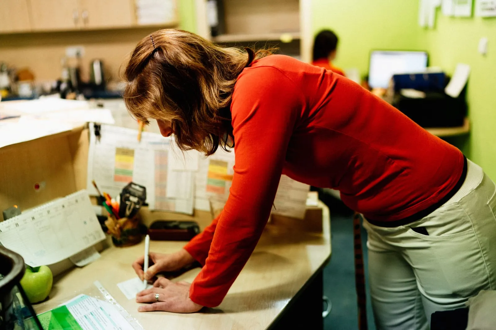 A woman in an orange top leans over a front desk to write a note, surrounded by schedules, a desk calendar and a computer in a bright green office.