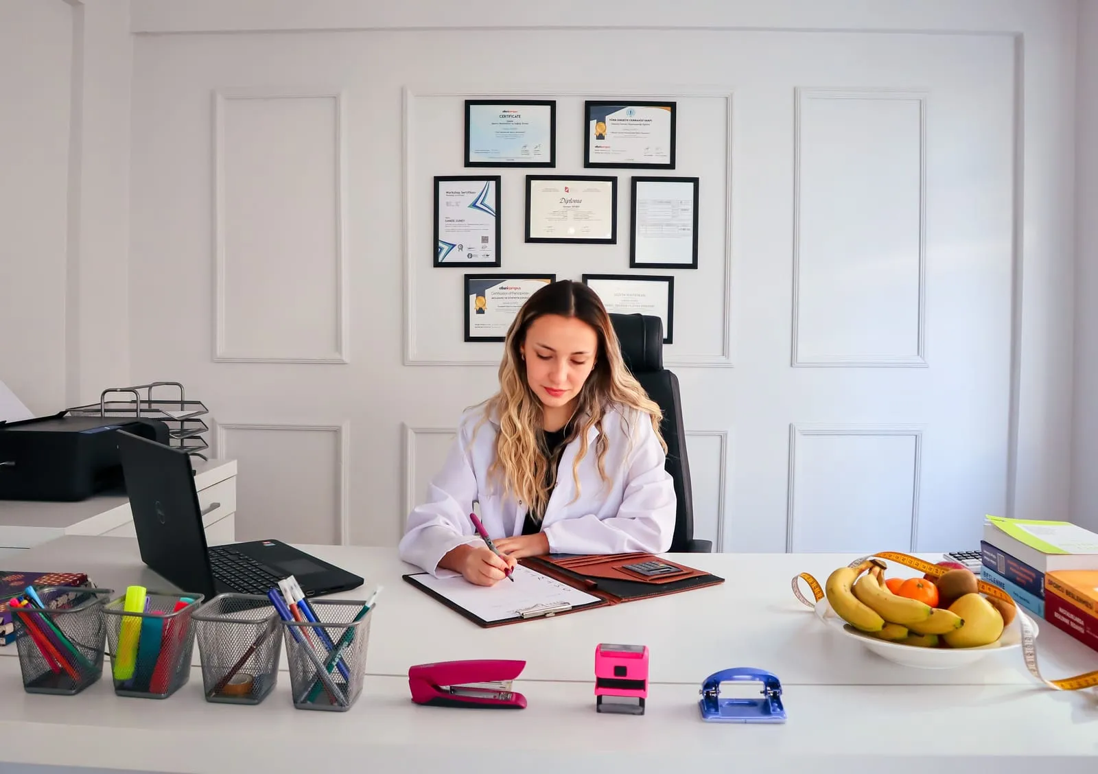 A woman in a white coat writes on a clipboard at a tidy white desk, with framed certificates on the wall behind her and a bowl of fruit nearby.