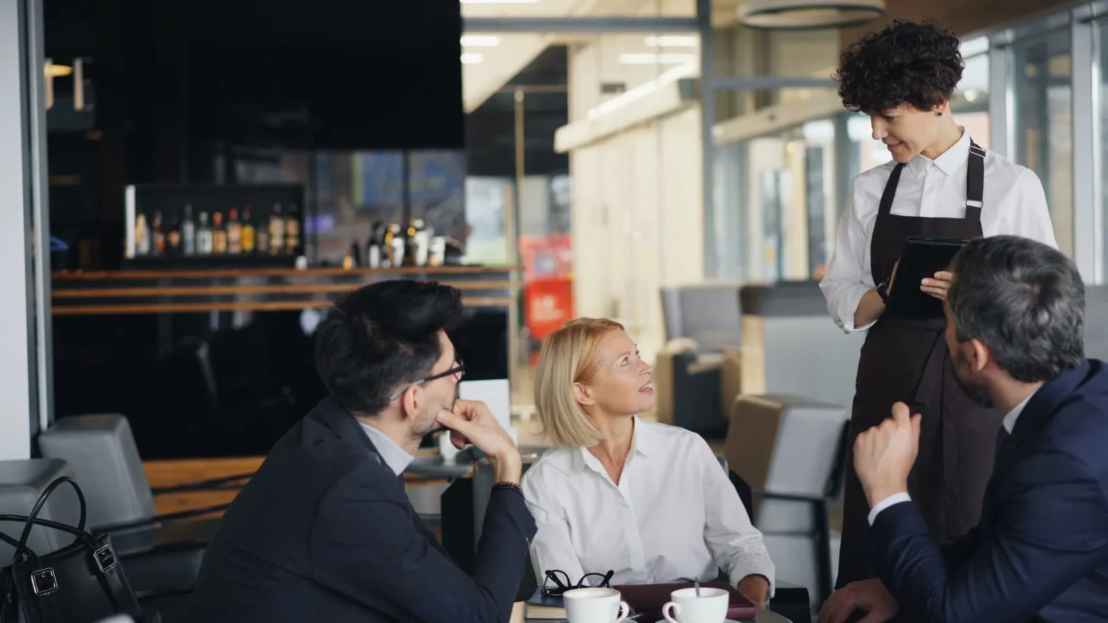 A server in an apron takes an order on a tablet from three seated business guests having coffee at a modern cafe table.
