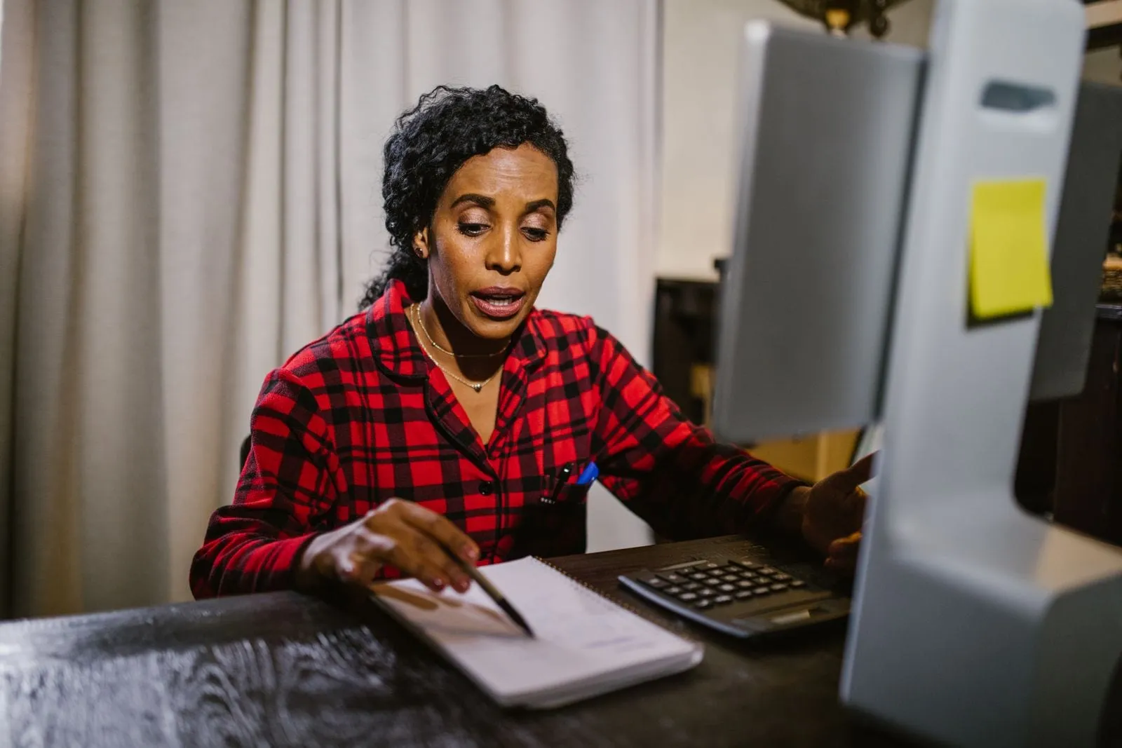 A woman at a home desk working through figures with a pen, a notebook and a calculator beside her computer