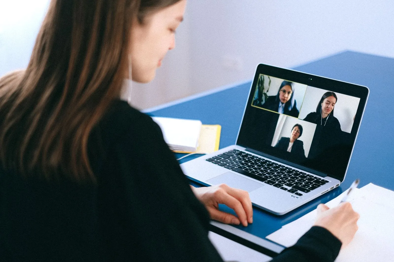 A student at a laptop on a video call with three people, taking notes on paper beside the keyboard