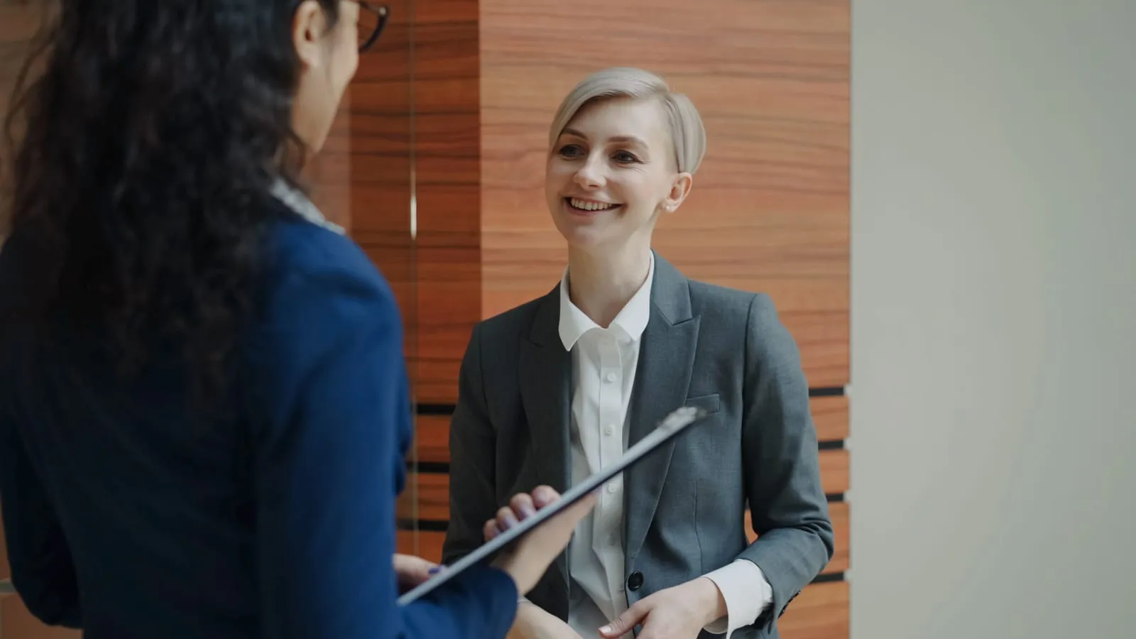 A smiling woman in a gray blazer chats with a colleague holding a clipboard in front of a wood-paneled wall.