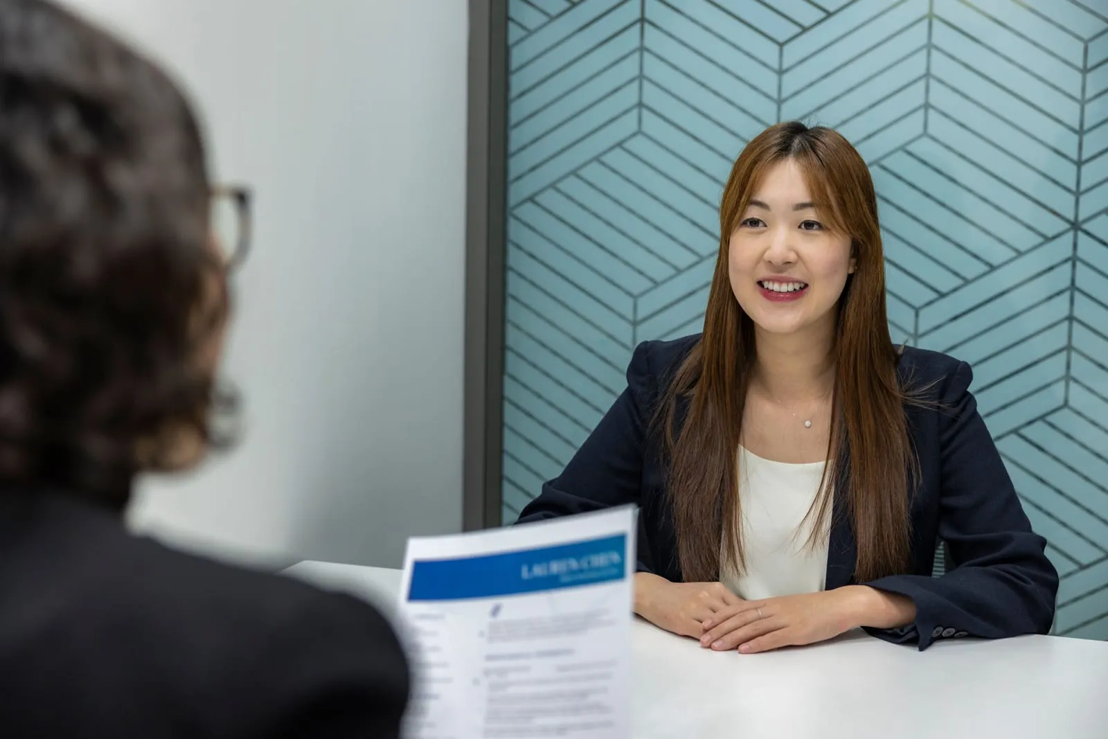 A smiling woman in a navy blazer sits across a white table during a job interview, while the interviewer in the foreground holds a printed resume.