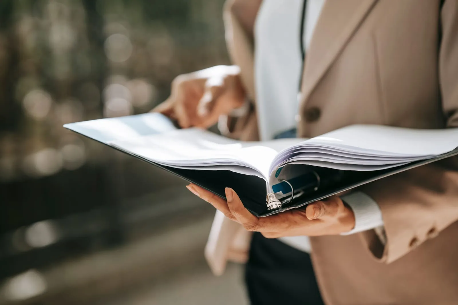 A person in a beige blazer holds an open black binder full of papers, turning a page, against a softly blurred outdoor background.