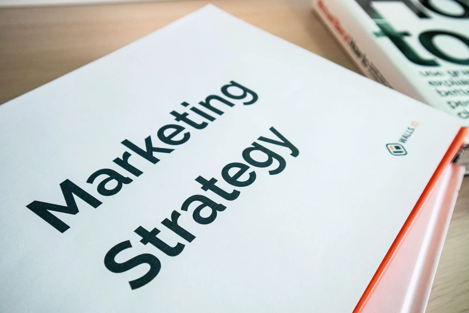 A printed Marketing Strategy cover page lies on a wooden desk beside an orange-edged binder and a book.