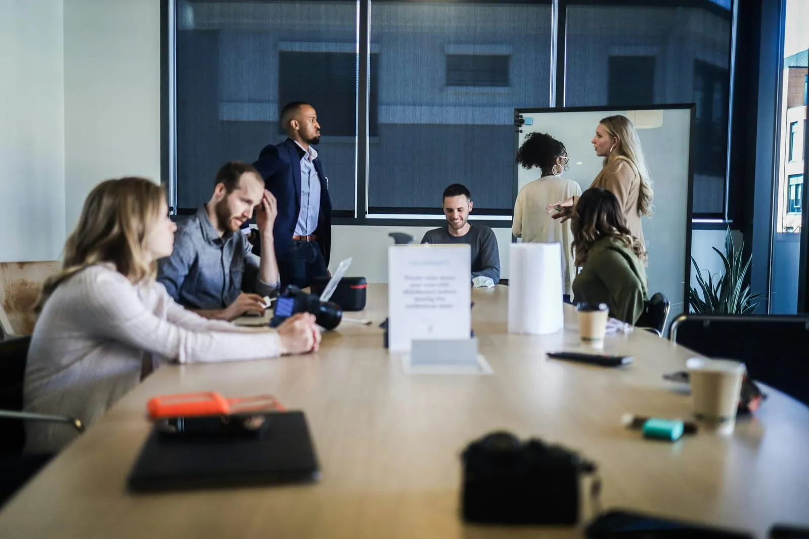 A team meets in a glass-walled conference room, some seated at a long table with laptops and a camera while others discuss ideas at a whiteboard.