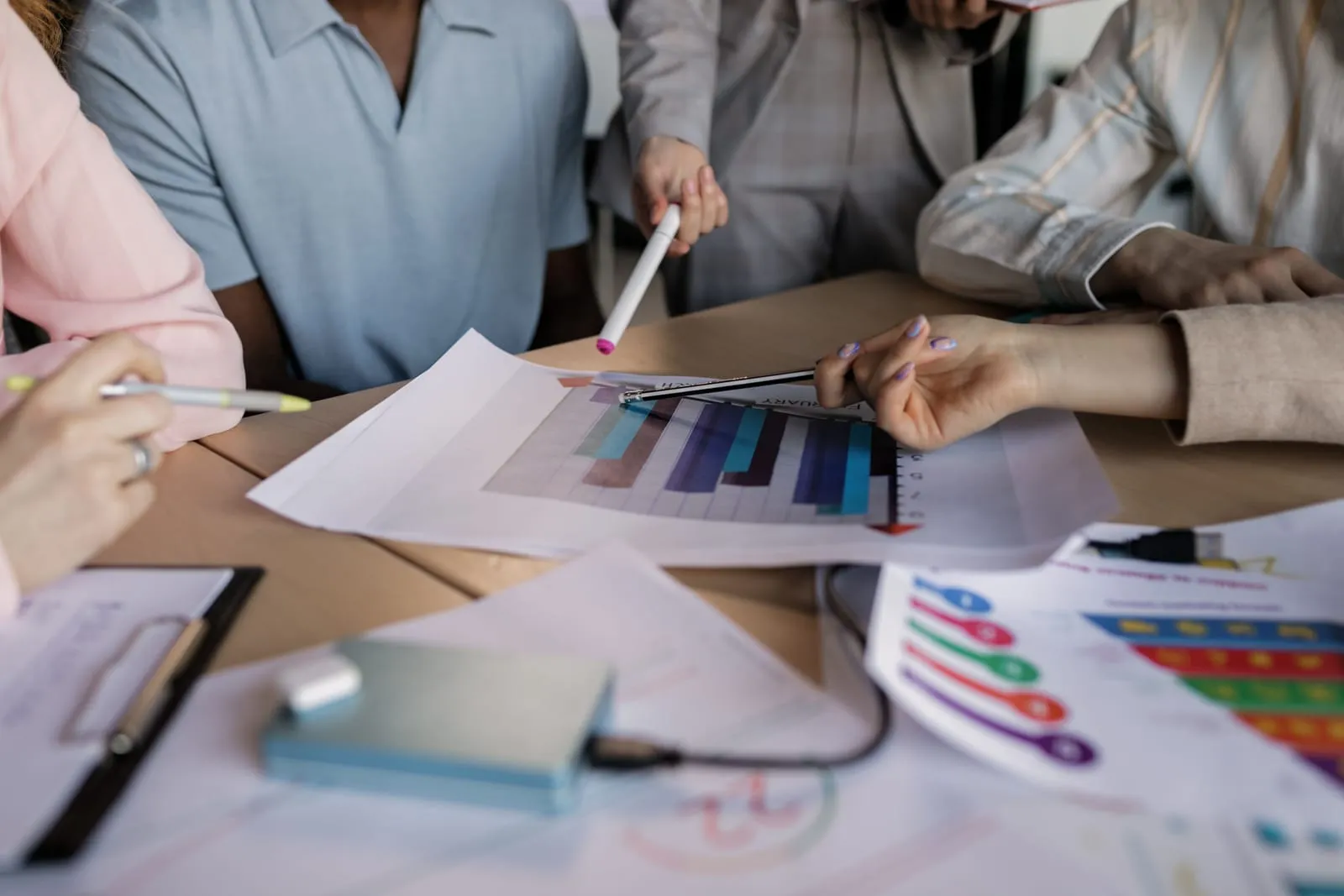 Colleagues gather around a table covered with printed bar charts and infographics, pointing at a graph with pens and markers.