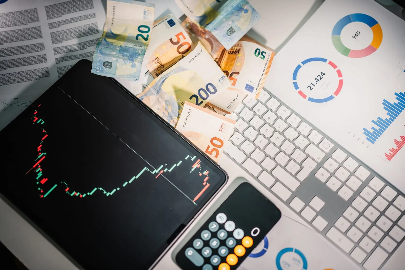 Overhead view of euro banknotes, a tablet with a candlestick price chart, a smartphone calculator, a keyboard and printed donut and bar charts.