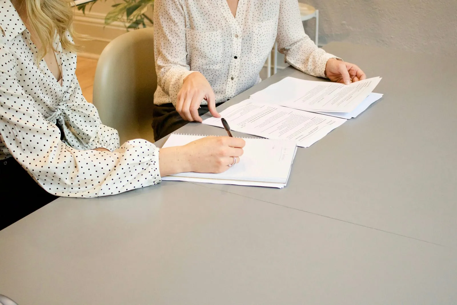 Two women in polka-dot blouses sit at a gray table, one writing in a notebook while the other reviews printed documents.