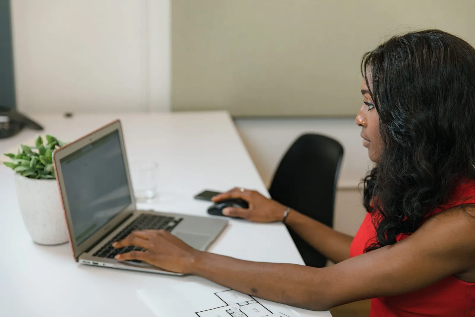 A woman in a red top works at a white desk, typing on a laptop and using a mouse, with a potted plant and floor plans nearby.