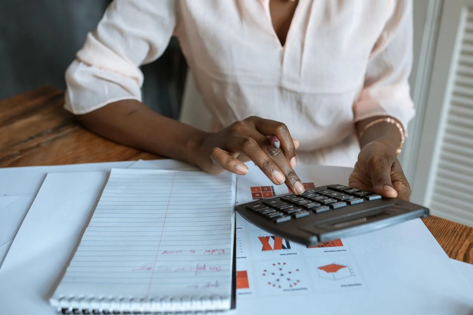 Close view of a woman pressing calculator keys at a desk with a lined notebook and printed charts