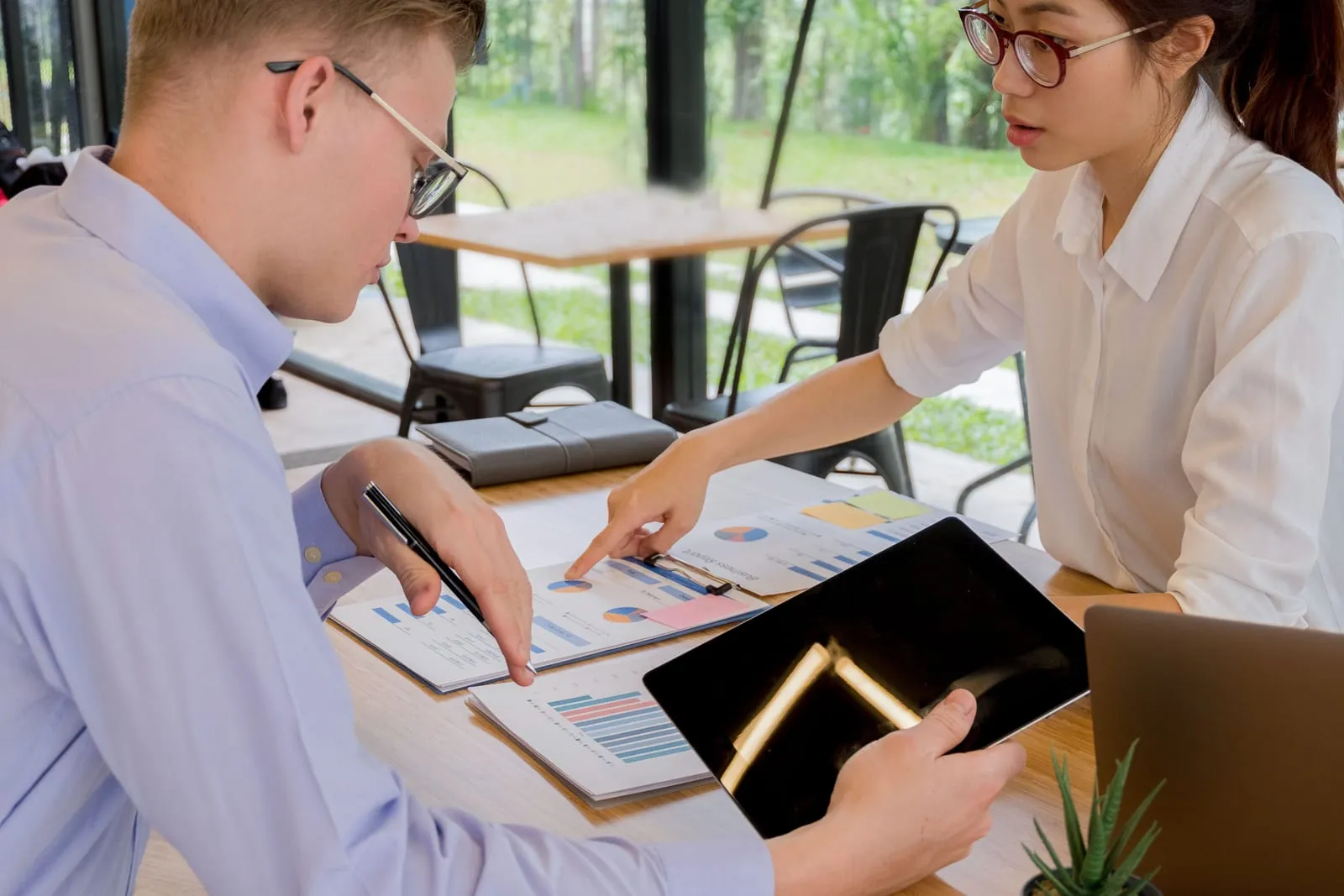 Two coworkers at a cafe-style table point at printed pie and bar charts, one holding a tablet, with greenery behind them