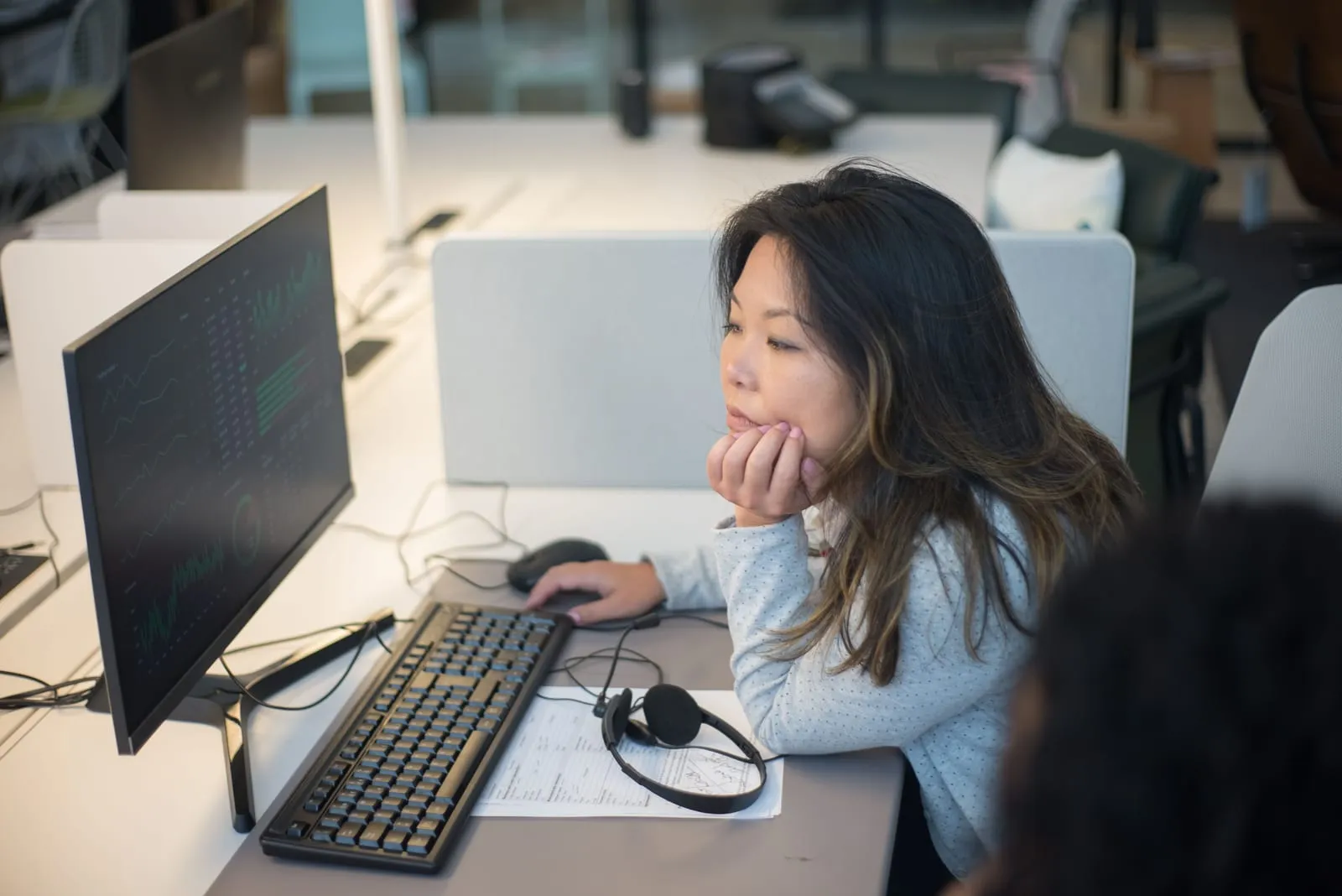 Woman rests her chin on her hand while studying a dark-themed data dashboard on a monitor at a shared desk, a headset beside the keyboard