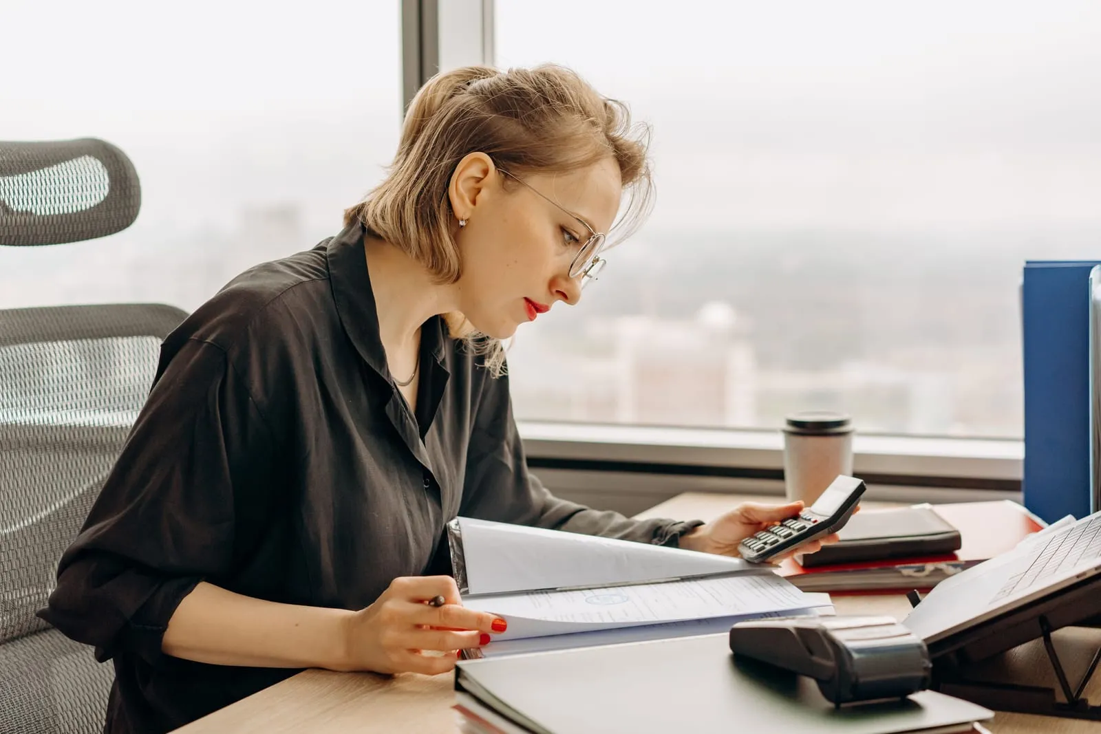 Woman in glasses reads through a binder of documents with a handheld calculator at a desk by a high-rise window