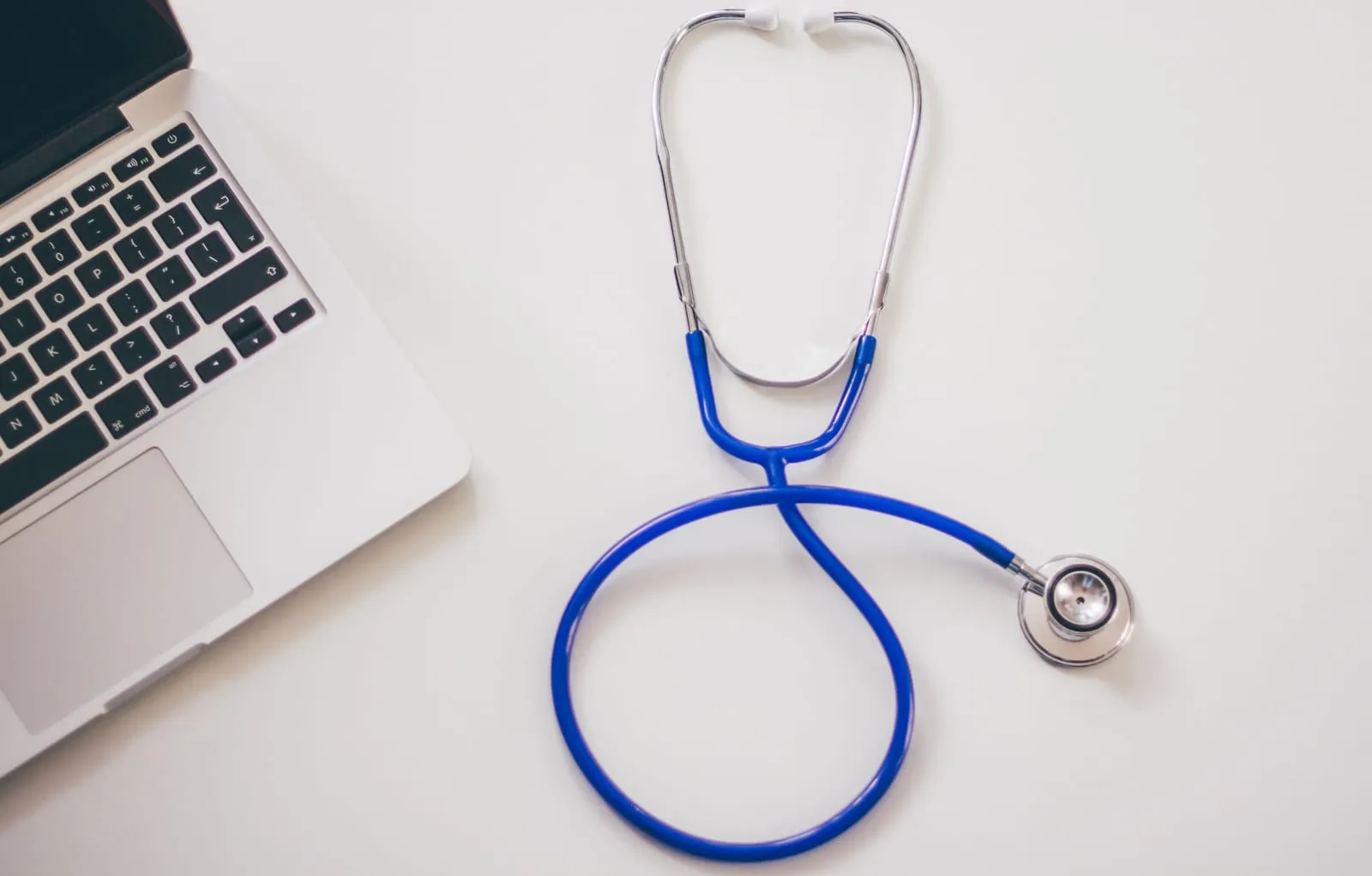 Blue stethoscope coiled on a white desk beside the corner of an open laptop, seen from above