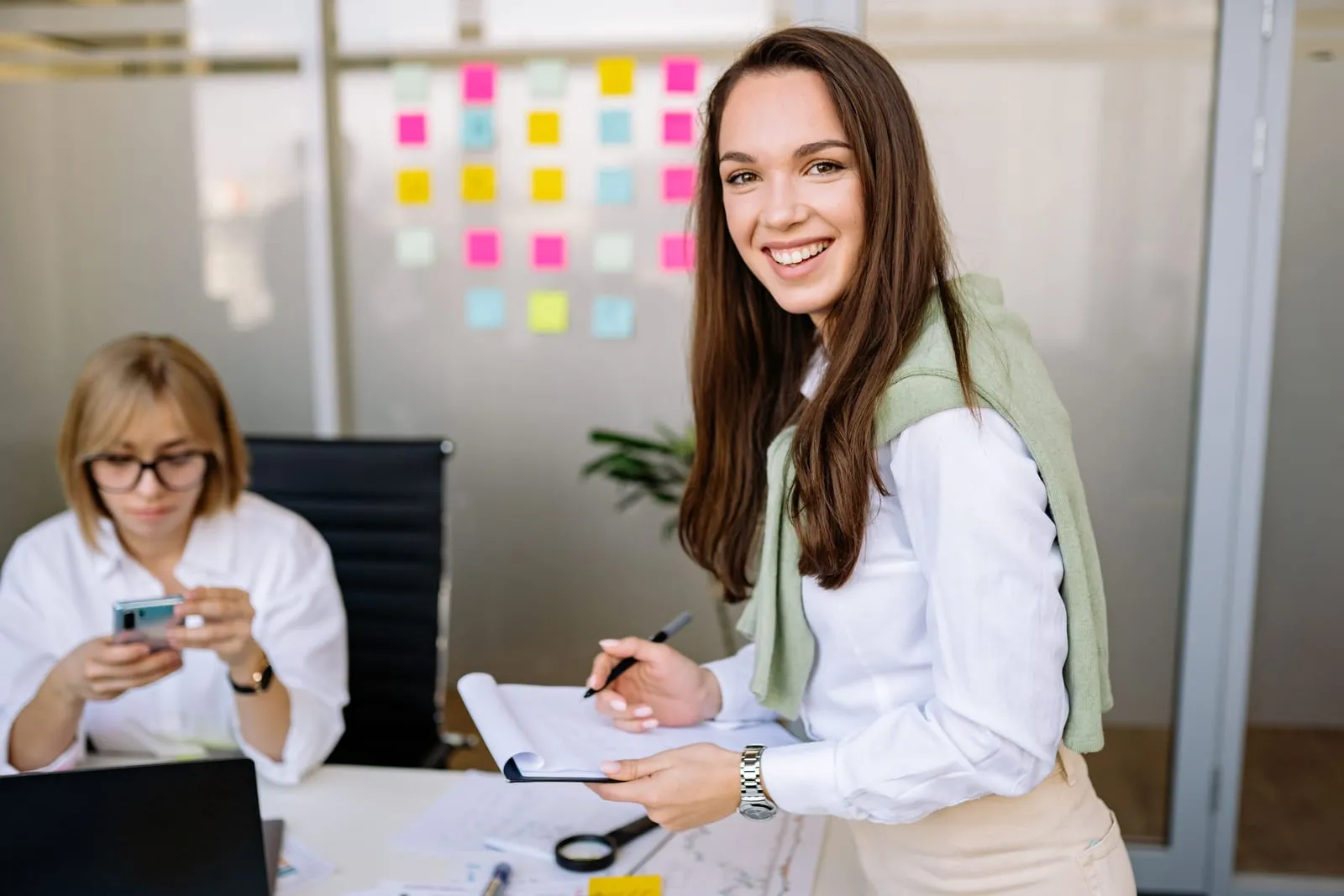 Smiling woman with a clipboard stands in an office with a wall of colorful sticky notes while a coworker checks her phone behind