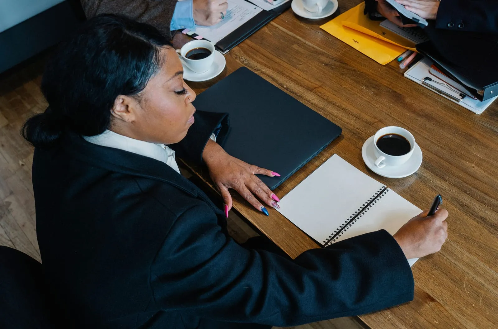 Woman in a dark suit writes in a spiral notebook at a wooden meeting table with coffee cups, a closed laptop and colleagues seen at the edges