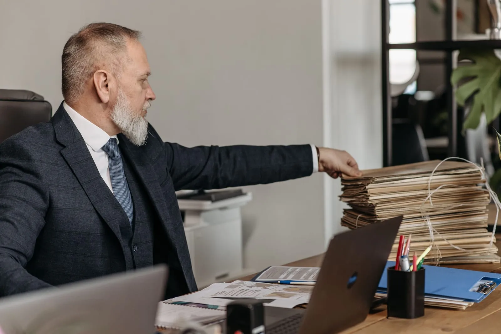 Gray-bearded man in a three-piece suit reaches toward a tall stack of paper files tied with string on a desk with a laptop