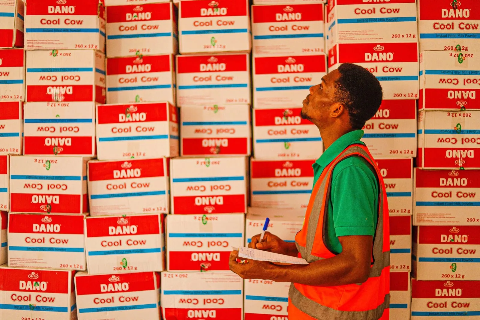 Warehouse worker in an orange safety vest looks up at a wall of stacked red and white milk powder cartons while holding a notepad