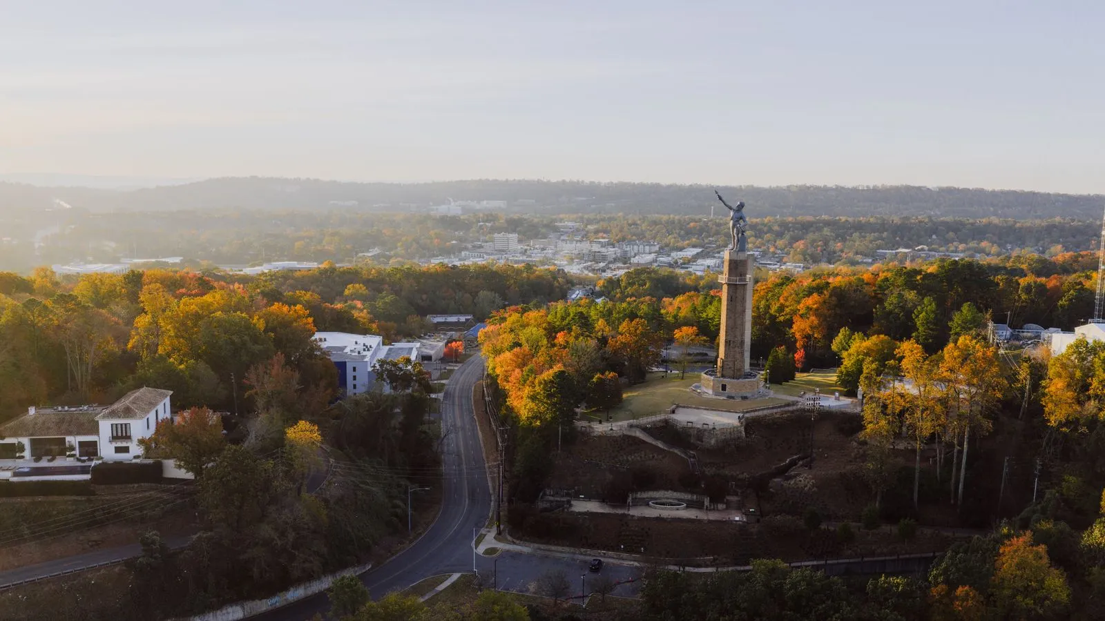 A tall statue on a stone column above autumn trees, a hazy city spreading out behind it