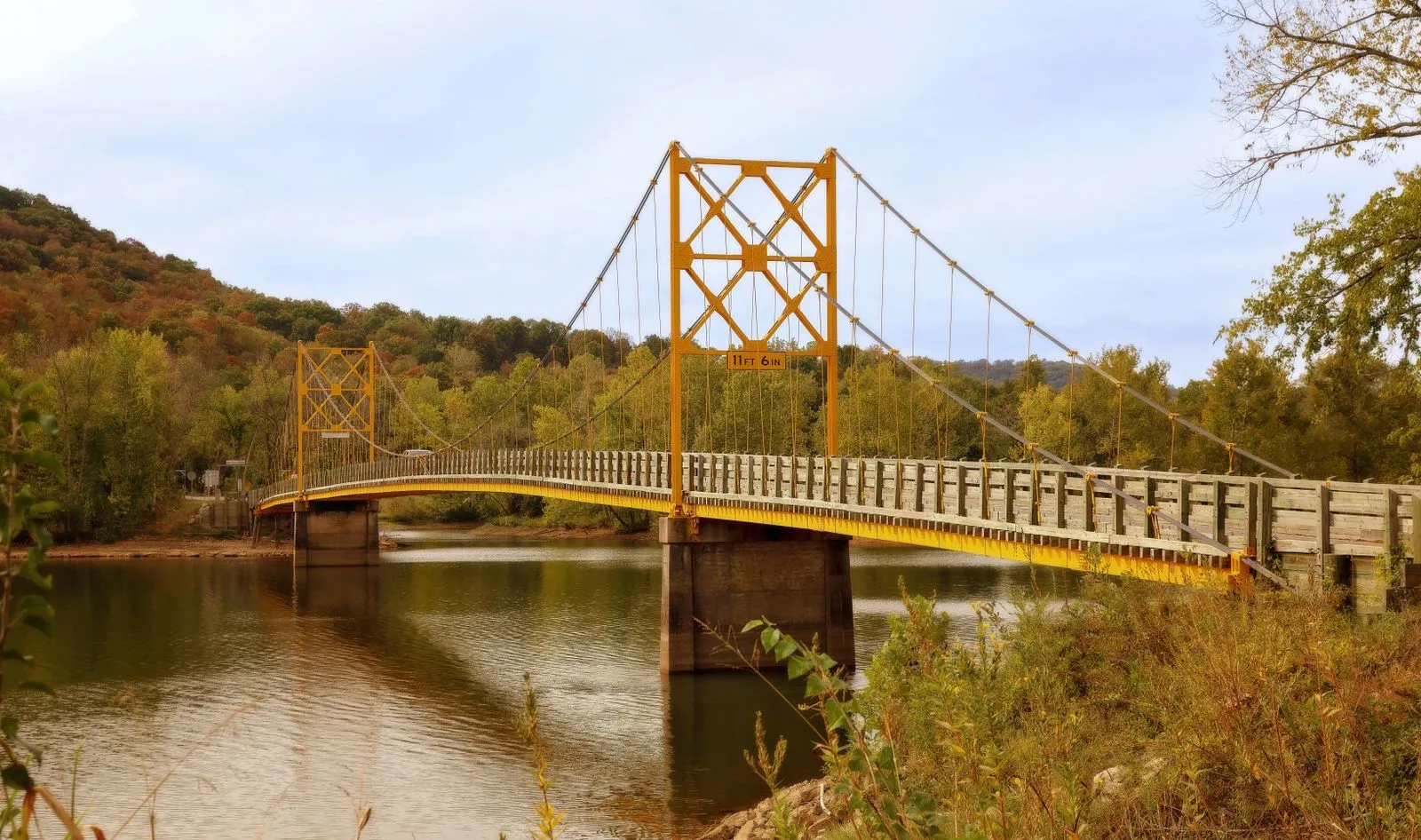 A yellow steel suspension bridge over a calm river between wooded autumn hills