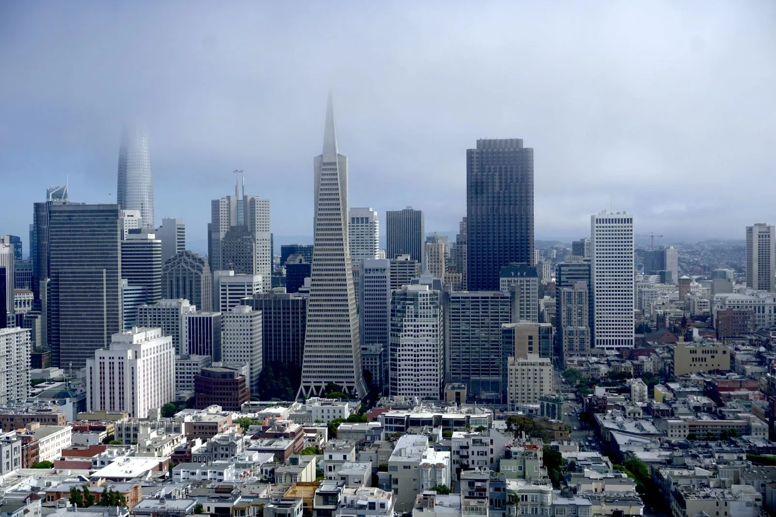 A dense downtown of towers around a tall pointed pyramid building, fog drifting over the tops