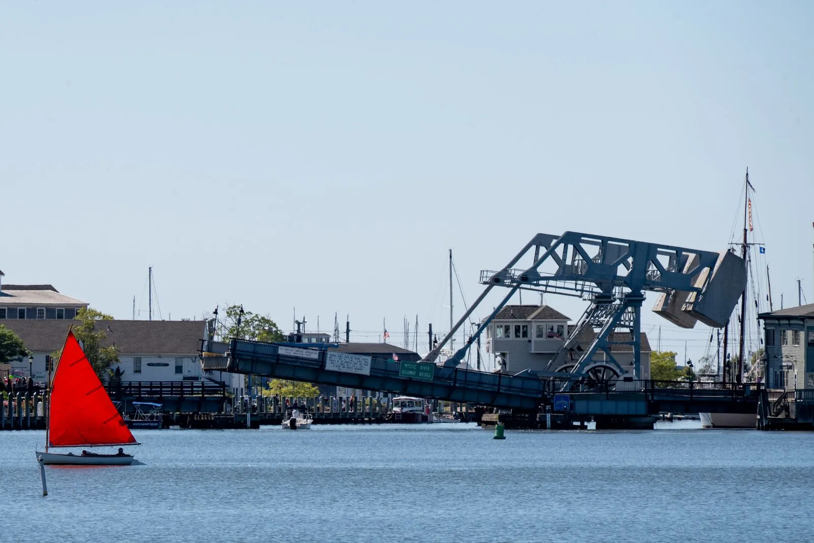 A red-sailed boat passing a raised steel drawbridge on a calm harbor