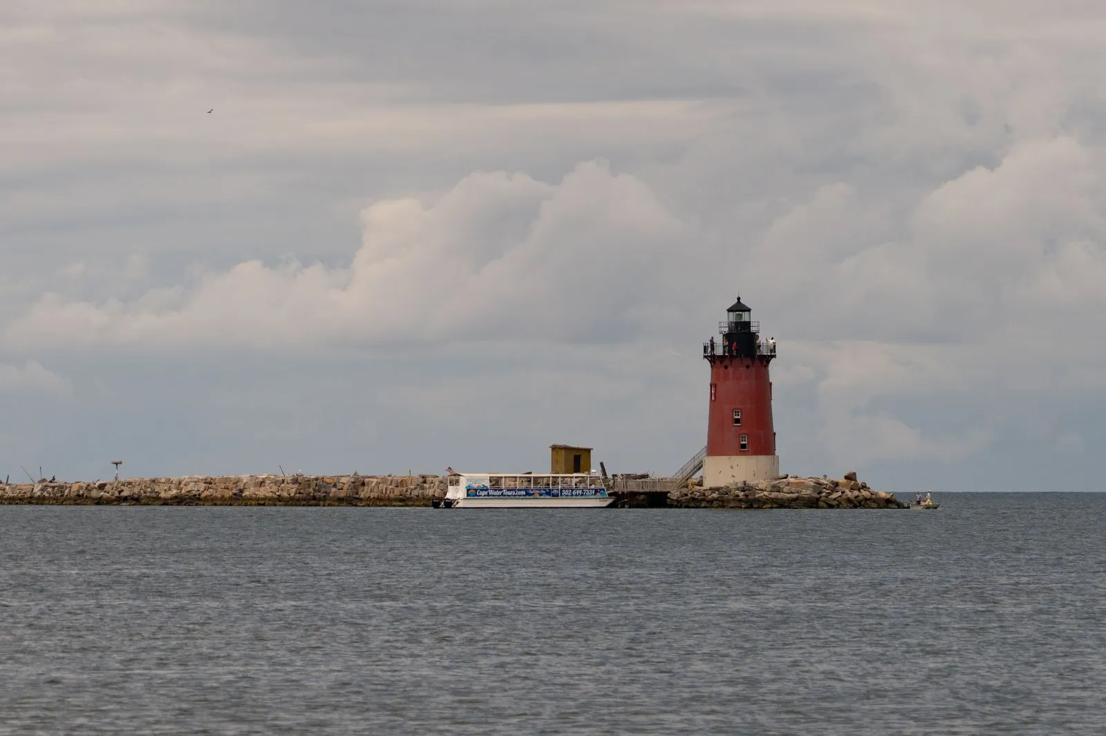 A red lighthouse at the end of a long stone breakwater in a wide gray bay