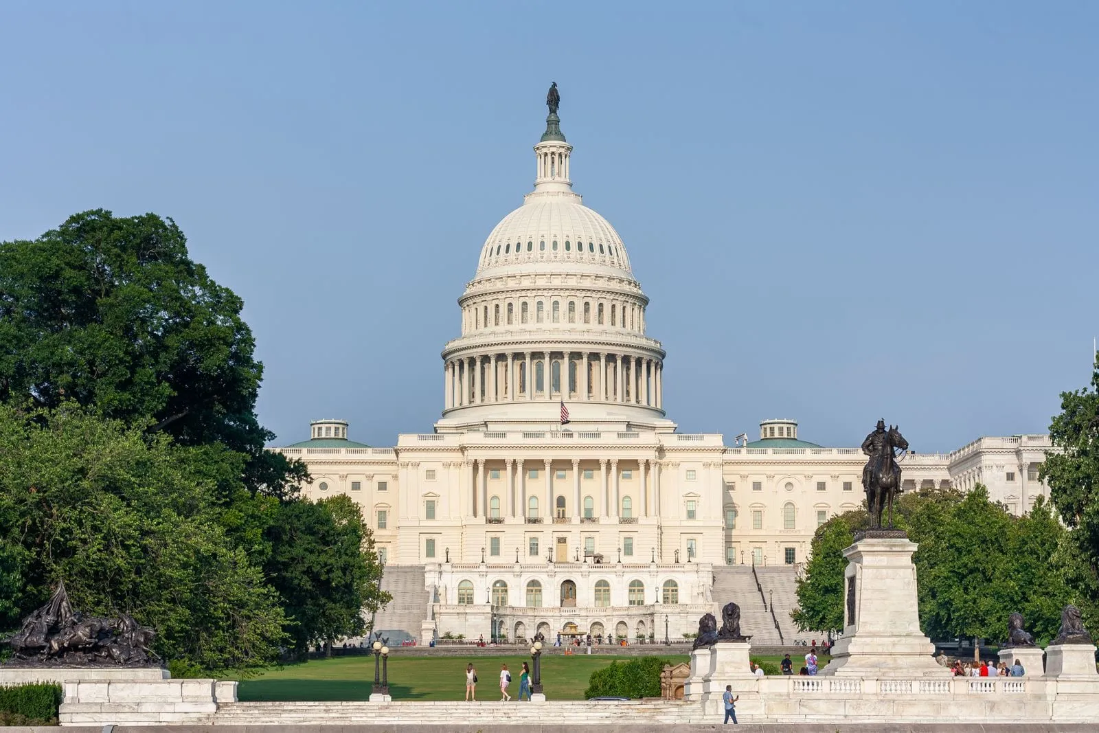 A white domed capitol building with a long colonnaded front behind a lawn, framed by trees and a bronze equestrian statue on a stone pedestal
