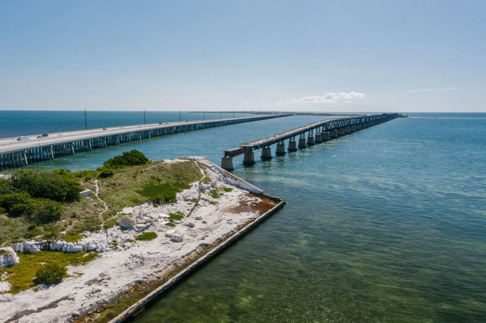 Aerial view of a long concrete bridge crossing shallow turquoise water beside the broken spans of an older bridge, a strip of scrub and pale rock in the foreground