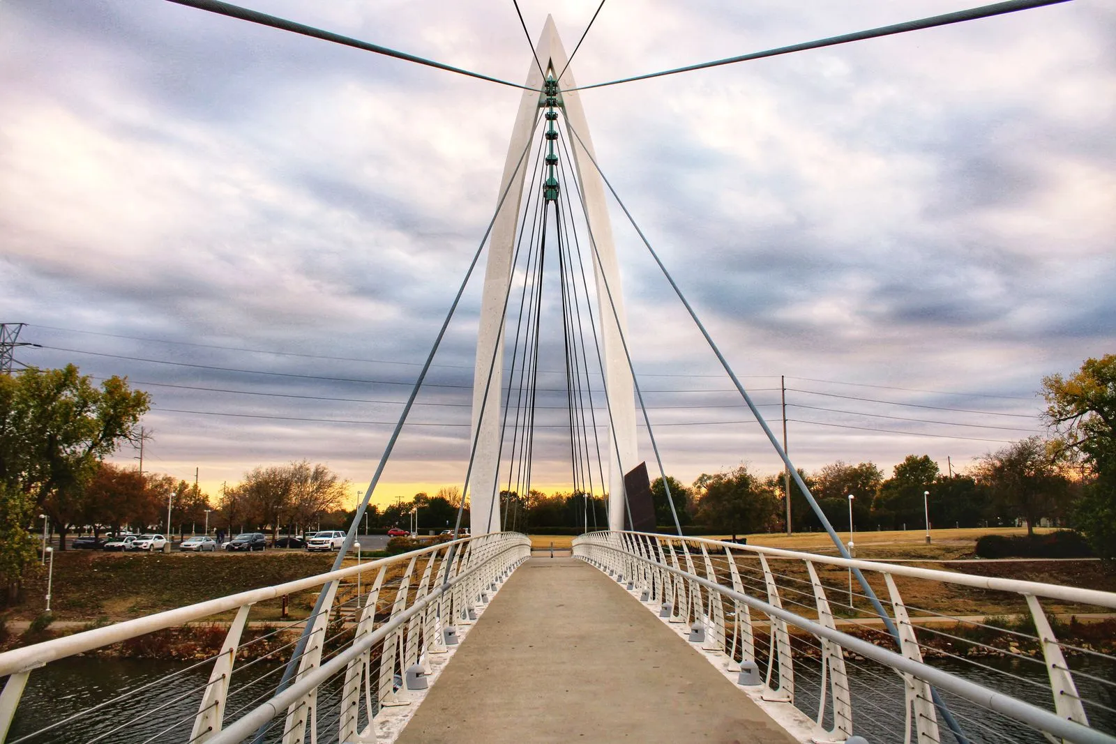 A white cable-stayed footbridge with a fan of cables crossing a river under a sunset sky