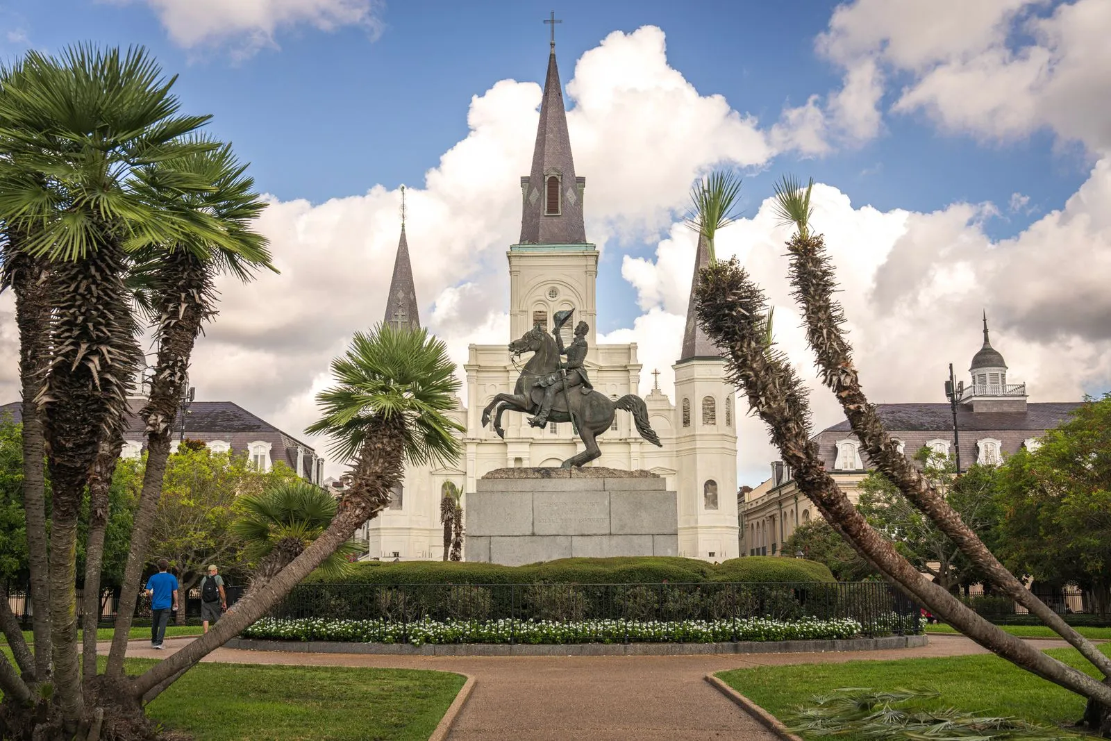 A white cathedral with three spires behind a bronze horseman statue, palms framing a garden
